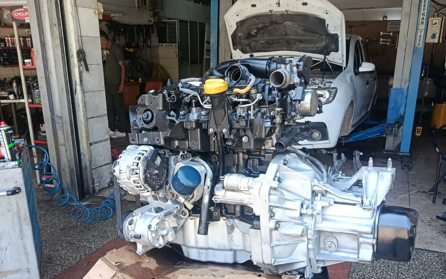 SE & FE Garaj tamirhanesinde tezgaha alınmış motor ve şanzıman takımı, arka planda lifte kaldırılmış kaputu açık araç