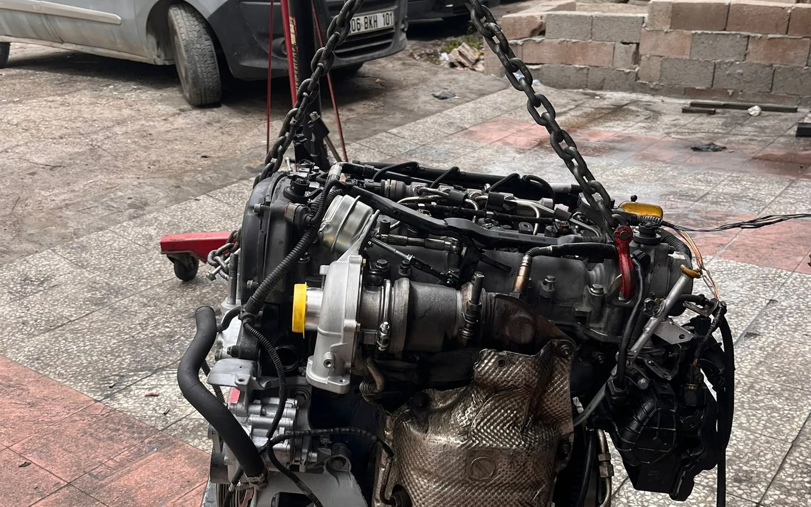 Egzoz sistemi görünür halde vinçle askıya alınmış motor, tamirhane önünde
